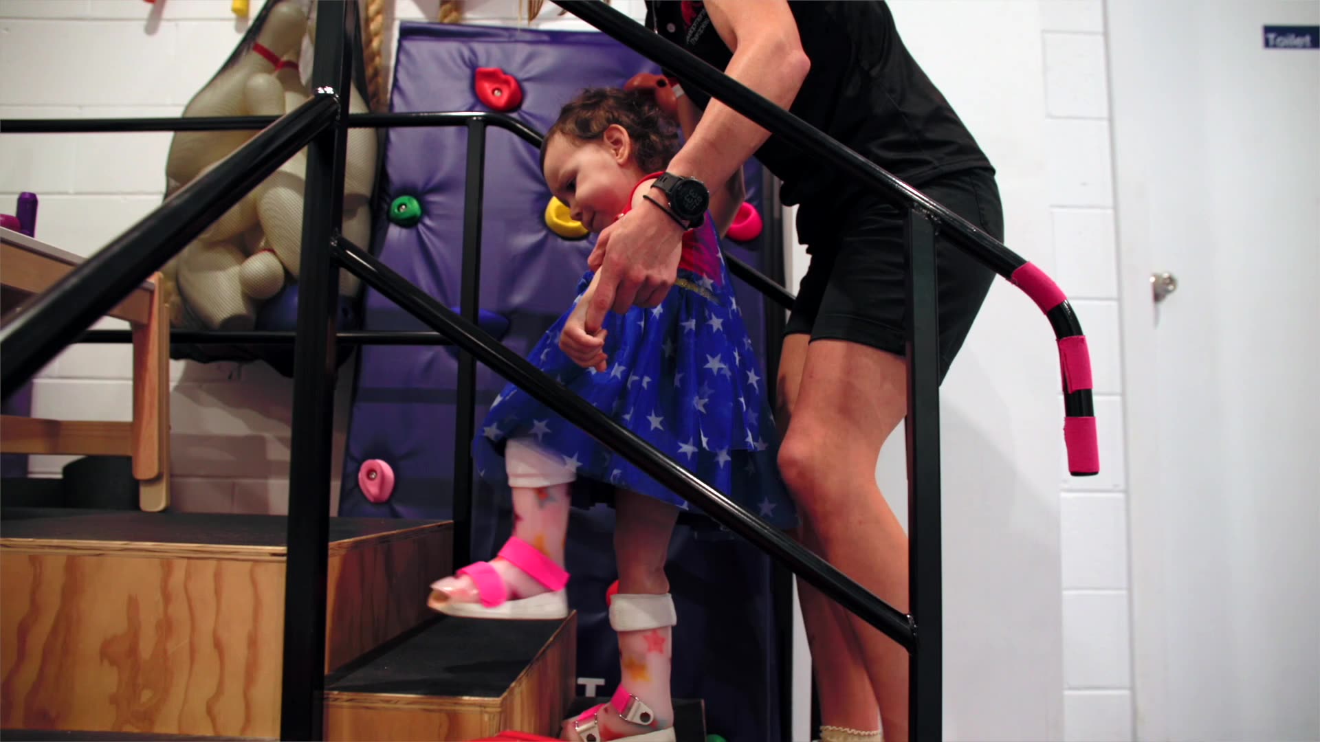 a toddler wearing orthotics is being guided up a set of practice stairs by a physiotherapist