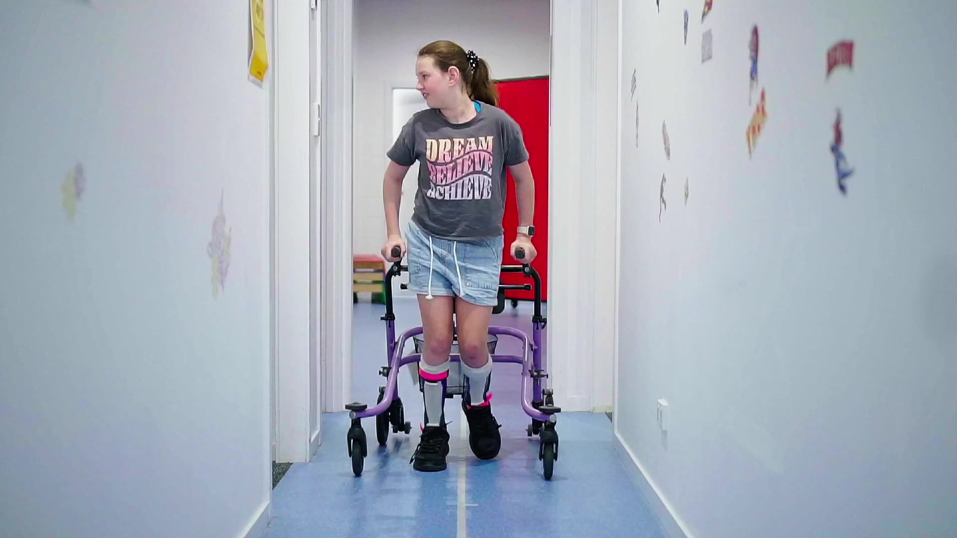 A girl walks down a corridor towards the camera using a walker and wear ankle foot orthotics AFO