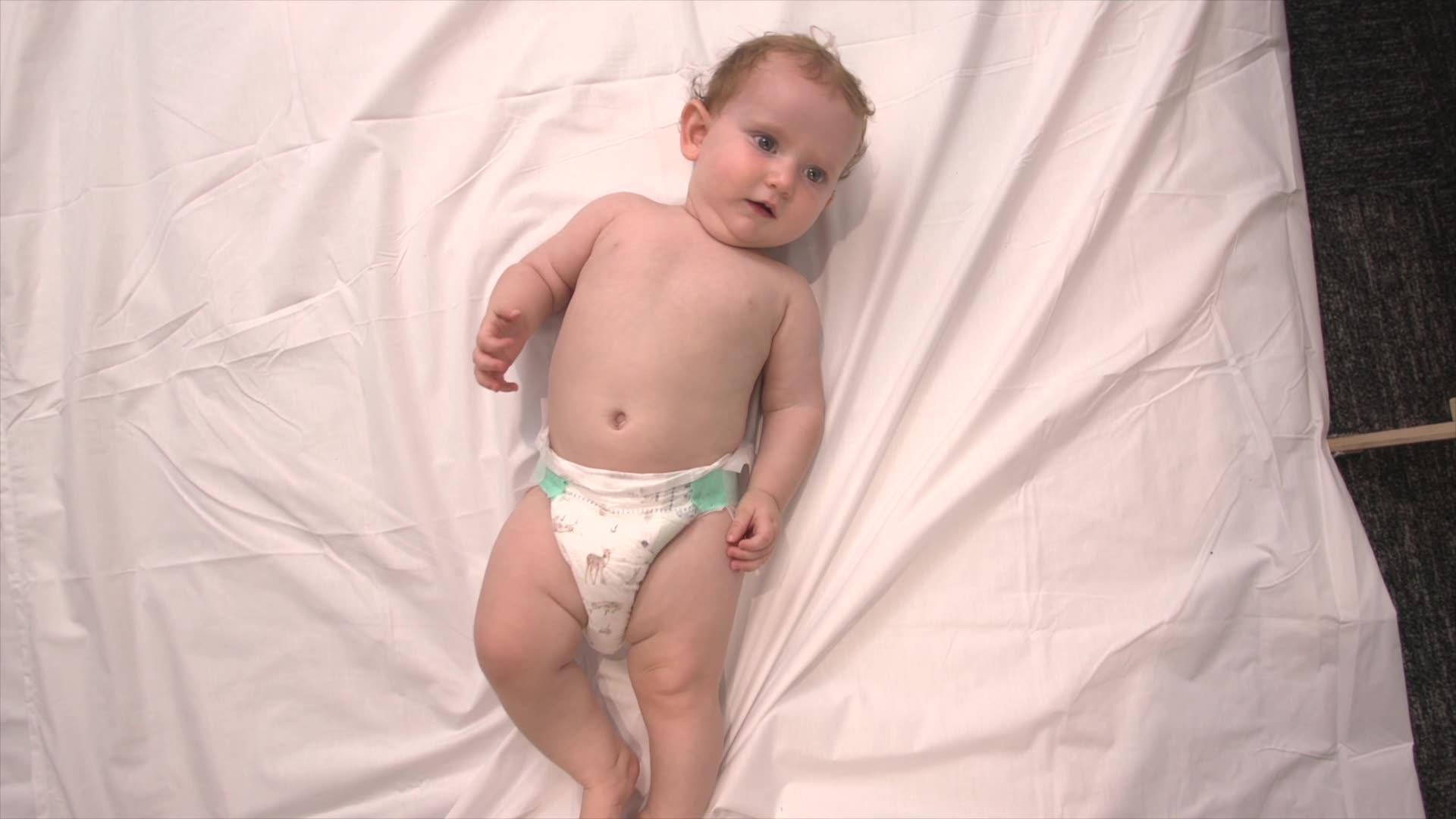 A baby in a nappy lies on it's back on a rug on the floor in a top down angle