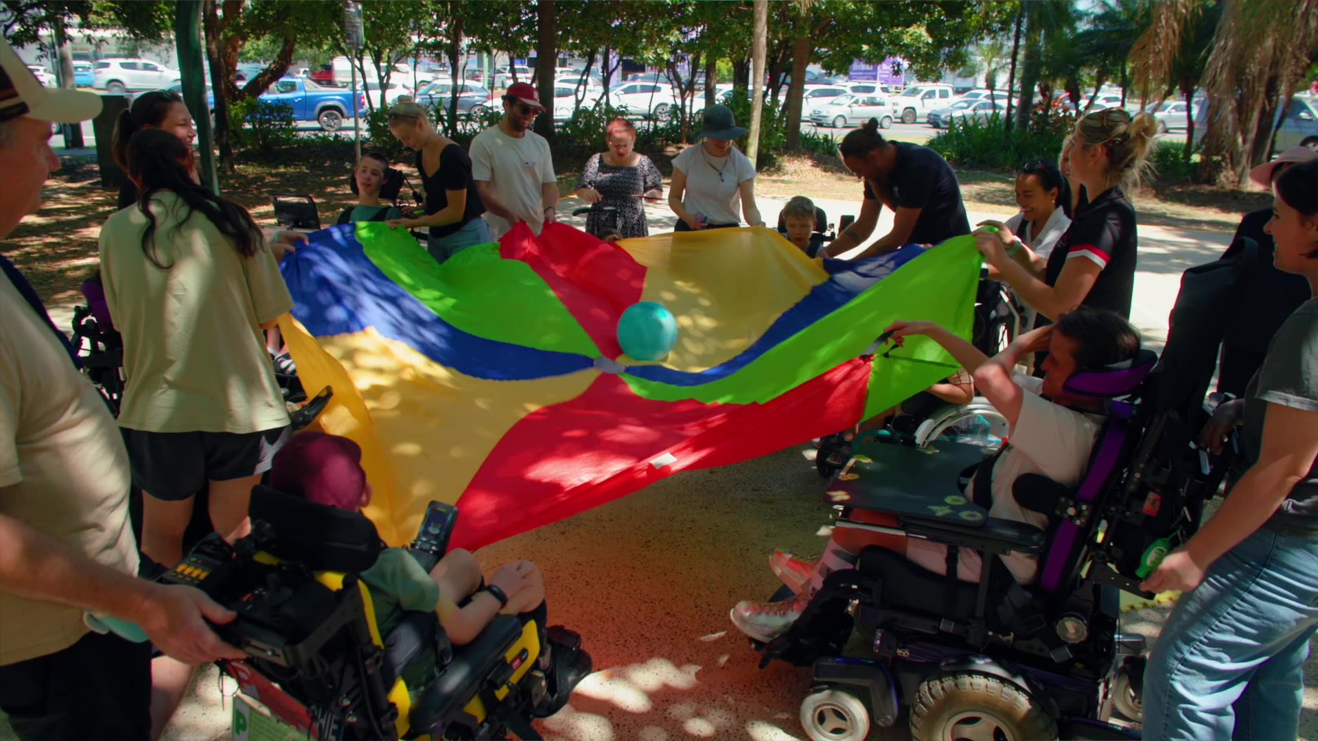 A top down view of a group of children in wheelchairs playing a parachute ball game with their carers and physiotherapists